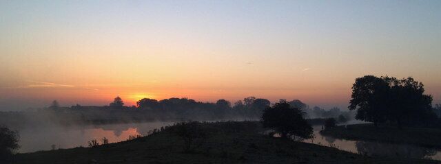 A sunrise over a misty landscape with a body of water and trees.