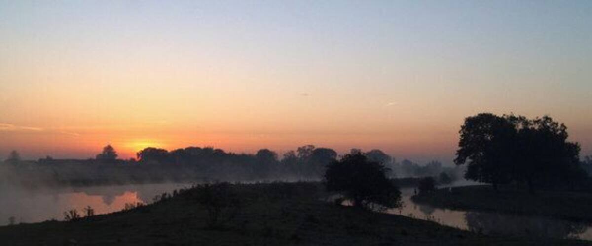 A sunrise over a misty landscape with a body of water and trees.