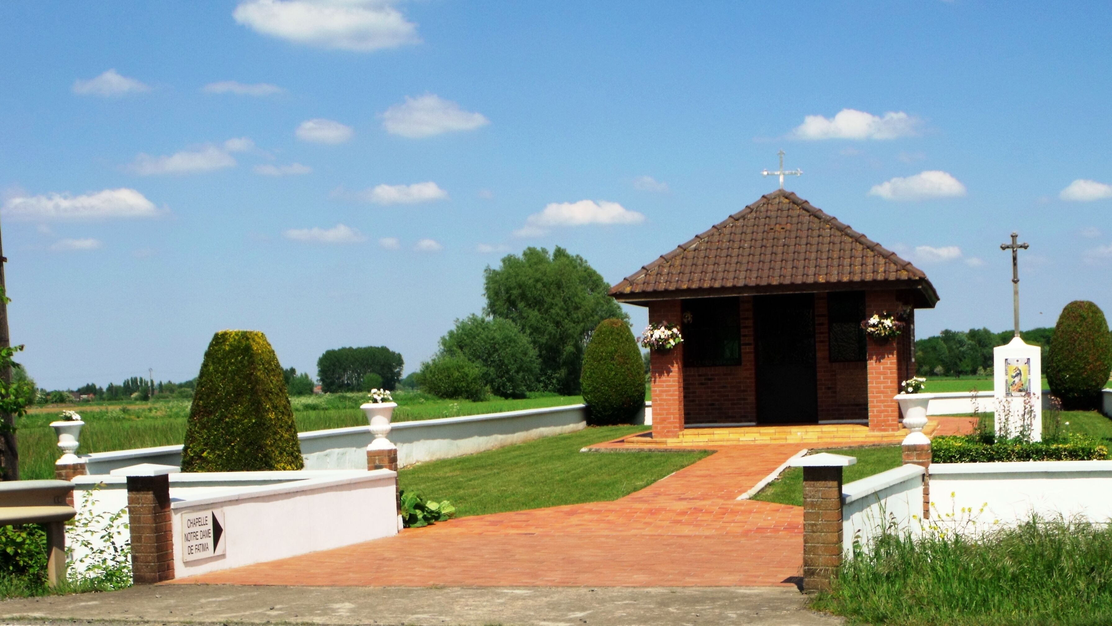 A small brick chapel with a tiled roof, surrounded by a white fence and a brick wall. There is a cross on top of the chapel and another on a nearby pole.
