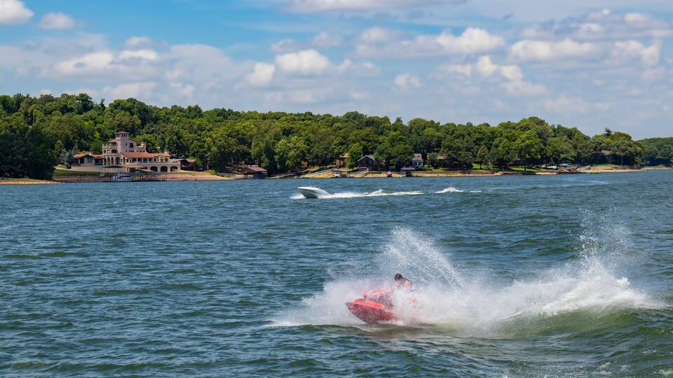 Person on a Person Watercraft plows through spray out on a lake wth another speedboat behind and beautiful homes with docks on the water among the trees on the shore under a cloudy sky