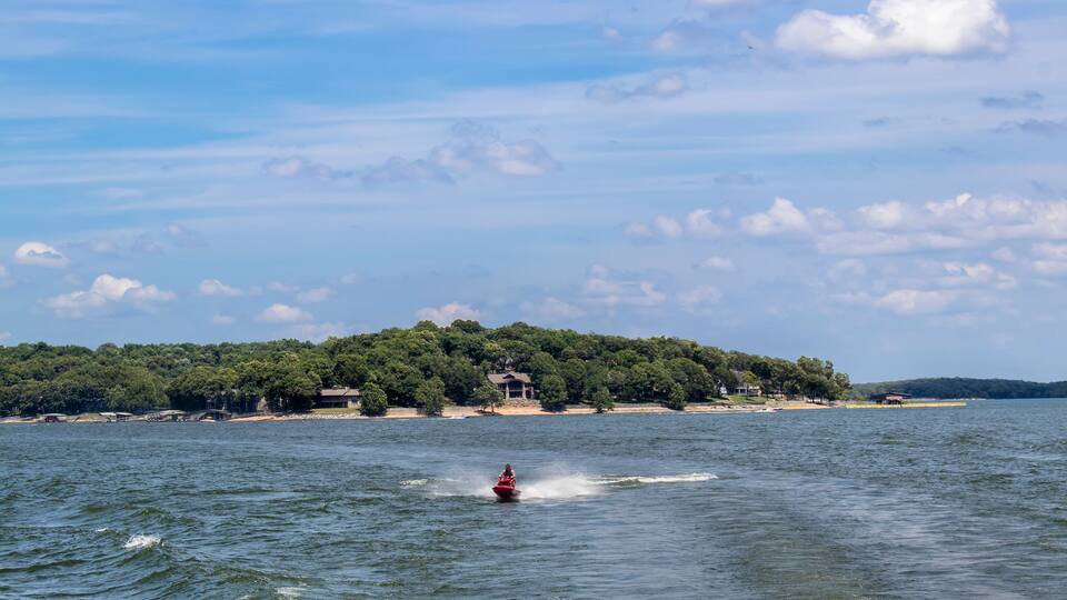 Person on red Personal Watercraft in choppy lake with homes and docks on shore and speedboat in background - Summer with cloudy blue cloud.