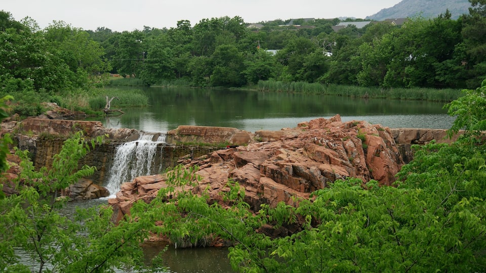 A waterfall cascading over red rocks into a calm lake surrounded by lush greenery and distant houses.