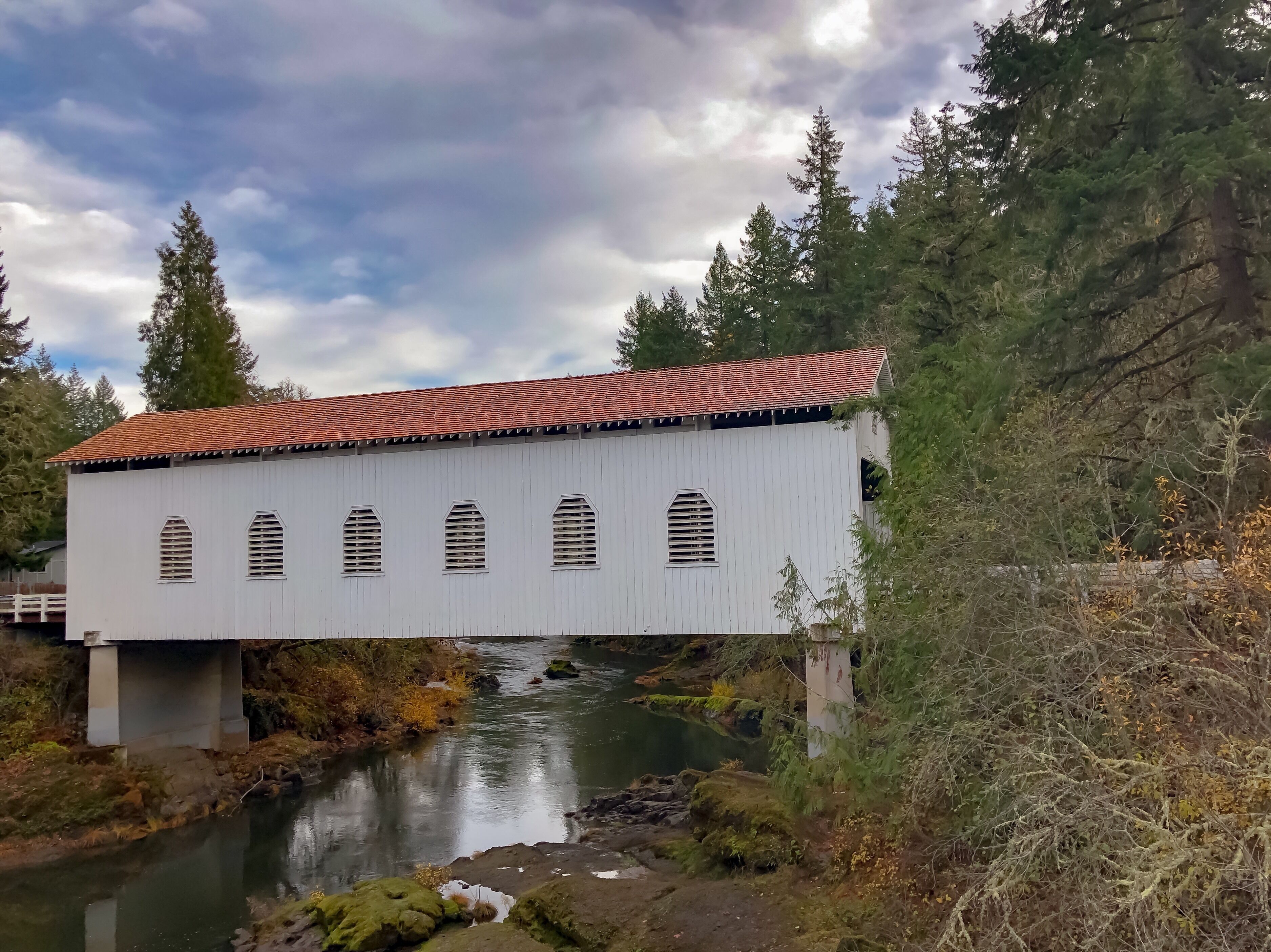A white covered bridge with a red roof spanning a river, surrounded by trees and autumn foliage.