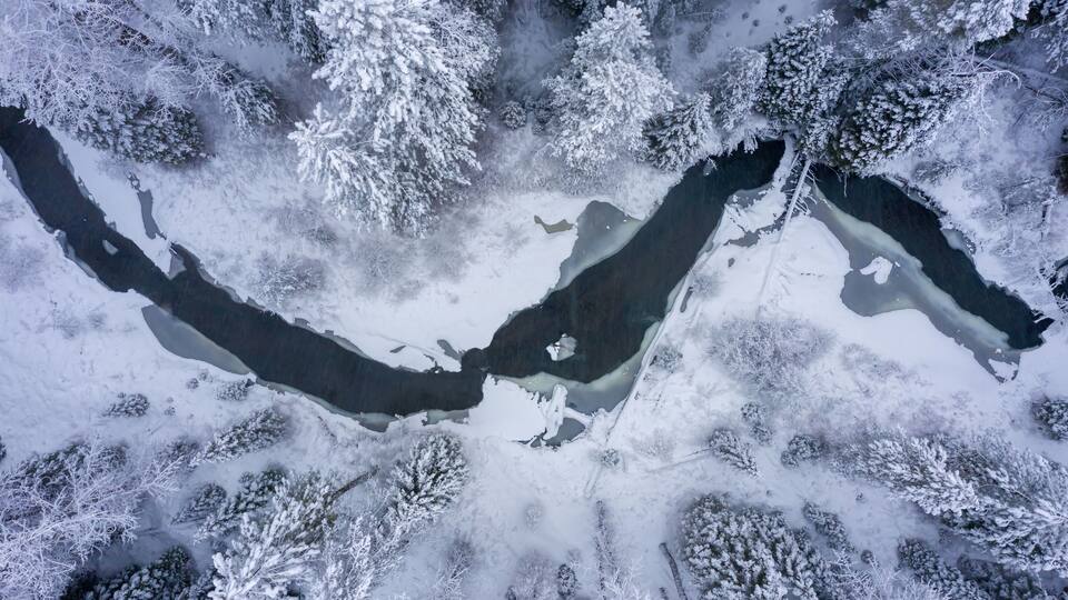 An aerial view of a snow-covered landscape with a frozen river and dense forest.