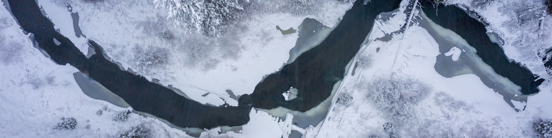 Aerial view of a partially frozen river flowing through a snow-covered forest in winter, creating a serene and picturesque landscape in Fort Klamath, Oregon, USA.