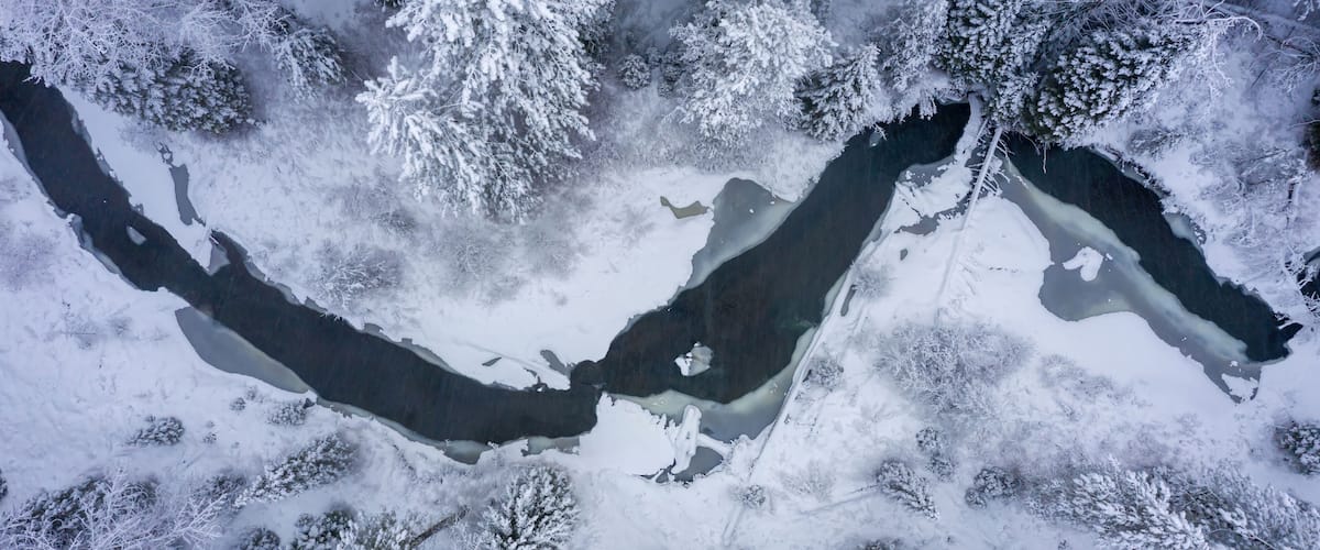 Aerial view of a partially frozen river flowing through a snow-covered forest in winter, creating a serene and picturesque landscape in Fort Klamath, Oregon, USA.