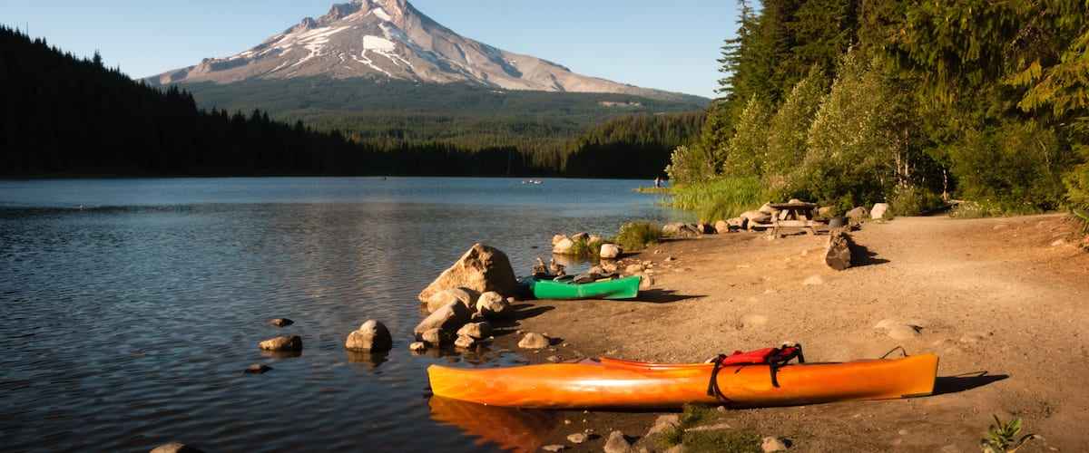A calm lake with kayaks on the shore, a mountain in the background, and dense forest on the right.