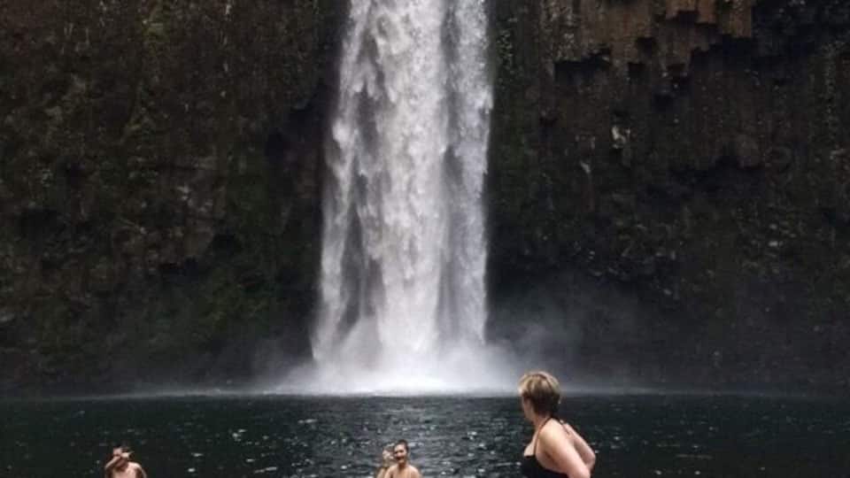 A waterfall in a forested area with three people standing in the water.