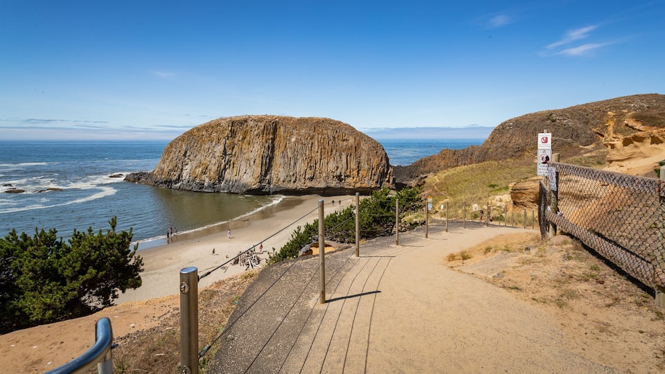 A coastal landscape with a prominent rock formation, a sandy beach, and a fenced pathway leading to the beach.