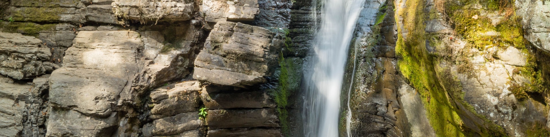 Une cascade d’eau se précipite le long d’une falaise rocheuse, entourée de pierres recouvertes de mousse.