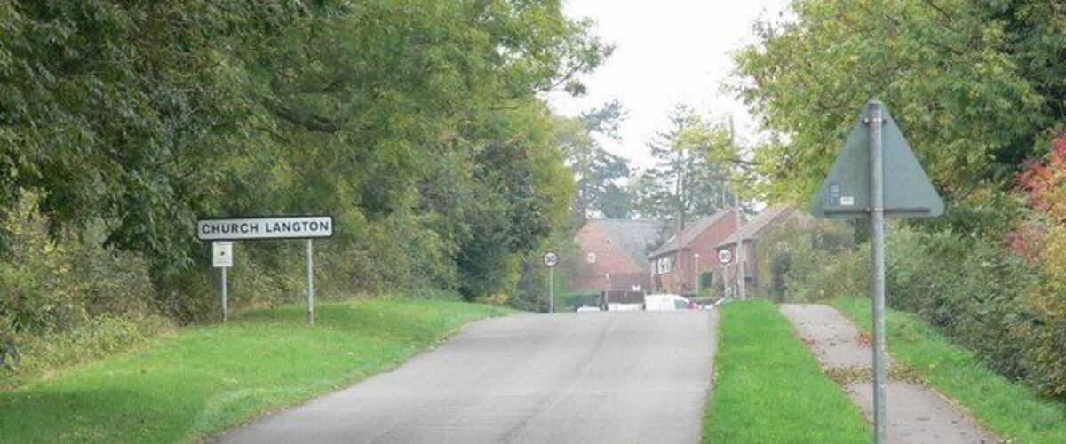 A rural road with a 'Church Langton' sign, a triangular warning sign, and a pedestrian path alongside.