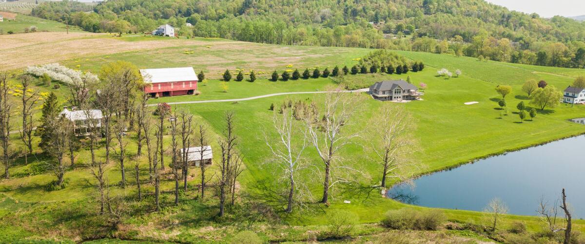 A rural landscape with a large house, a barn, a pond, and rolling hills.