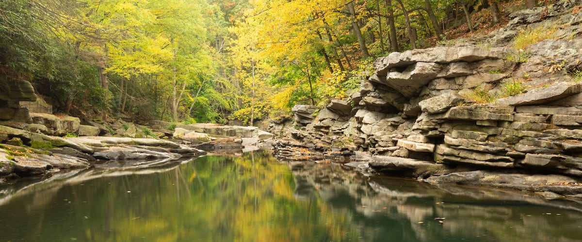 A calm river reflecting yellow autumn leaves, surrounded by rocky terrain and dense trees.