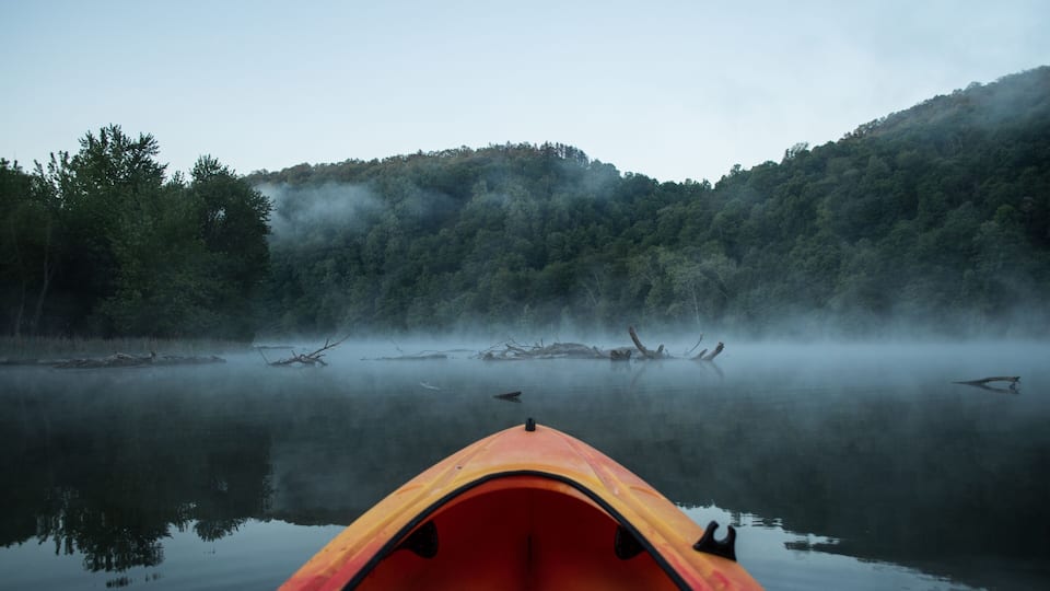 A red kayak on calm water with misty forested hills in the background.
