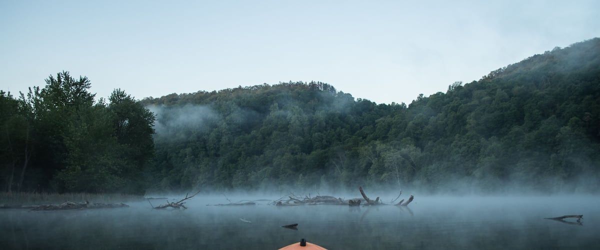 A red kayak on calm water with misty forested hills in the background.