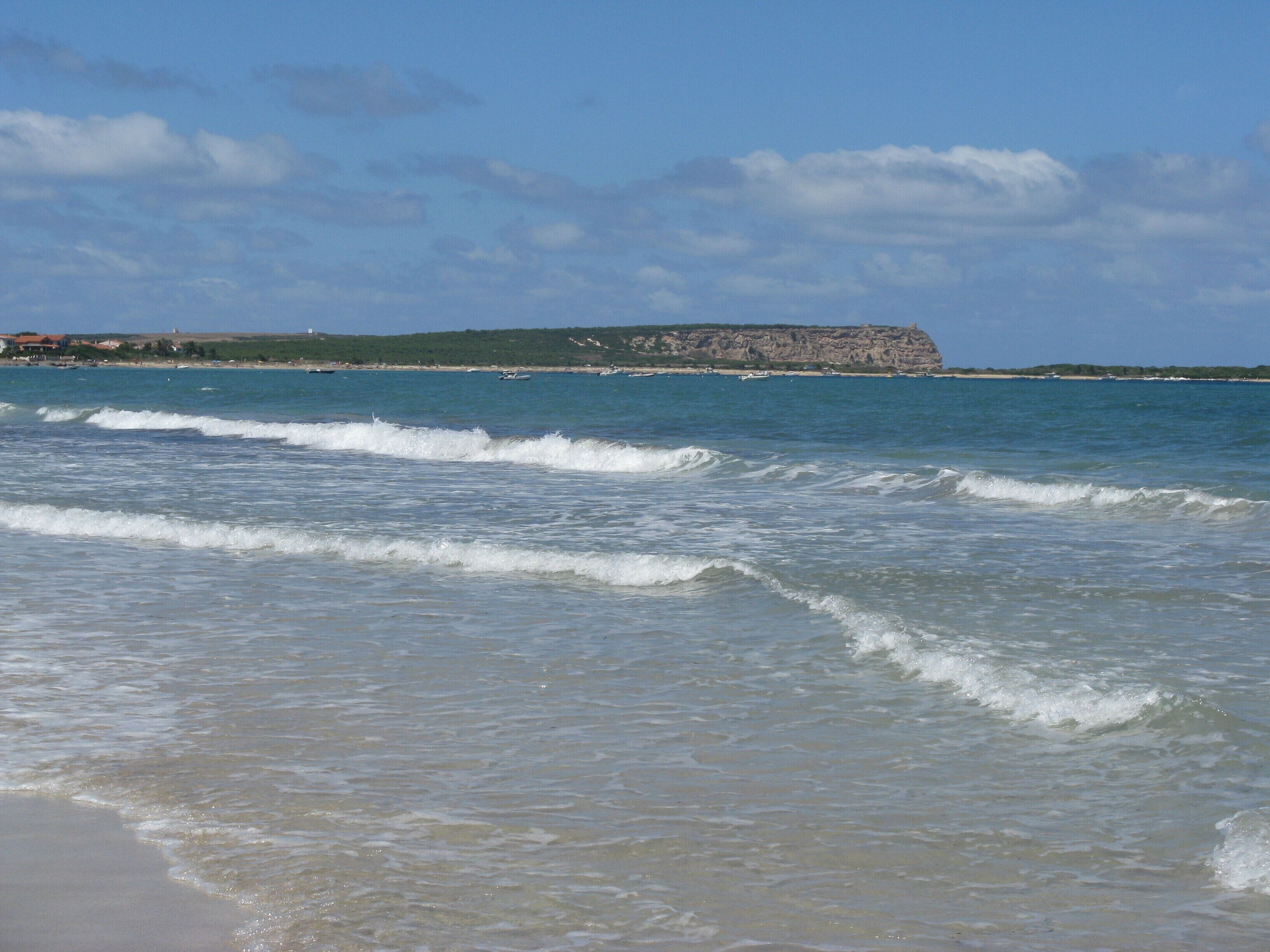 A beach with waves, a distant rocky cliff, and a clear sky.