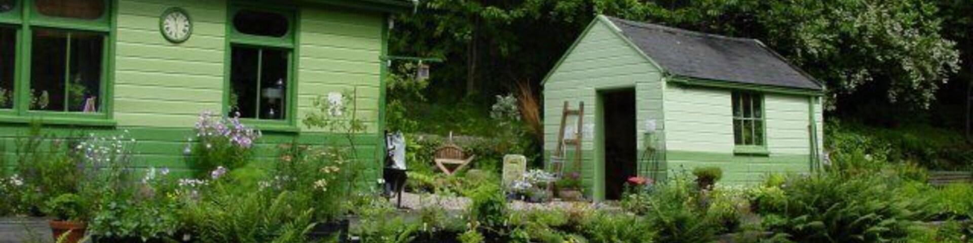A green house with a clock, surrounded by a garden with various plants and flowers.