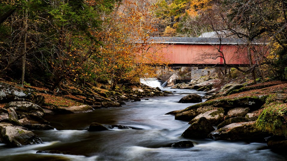 A red covered bridge over a flowing river, surrounded by autumn foliage and rocky terrain.