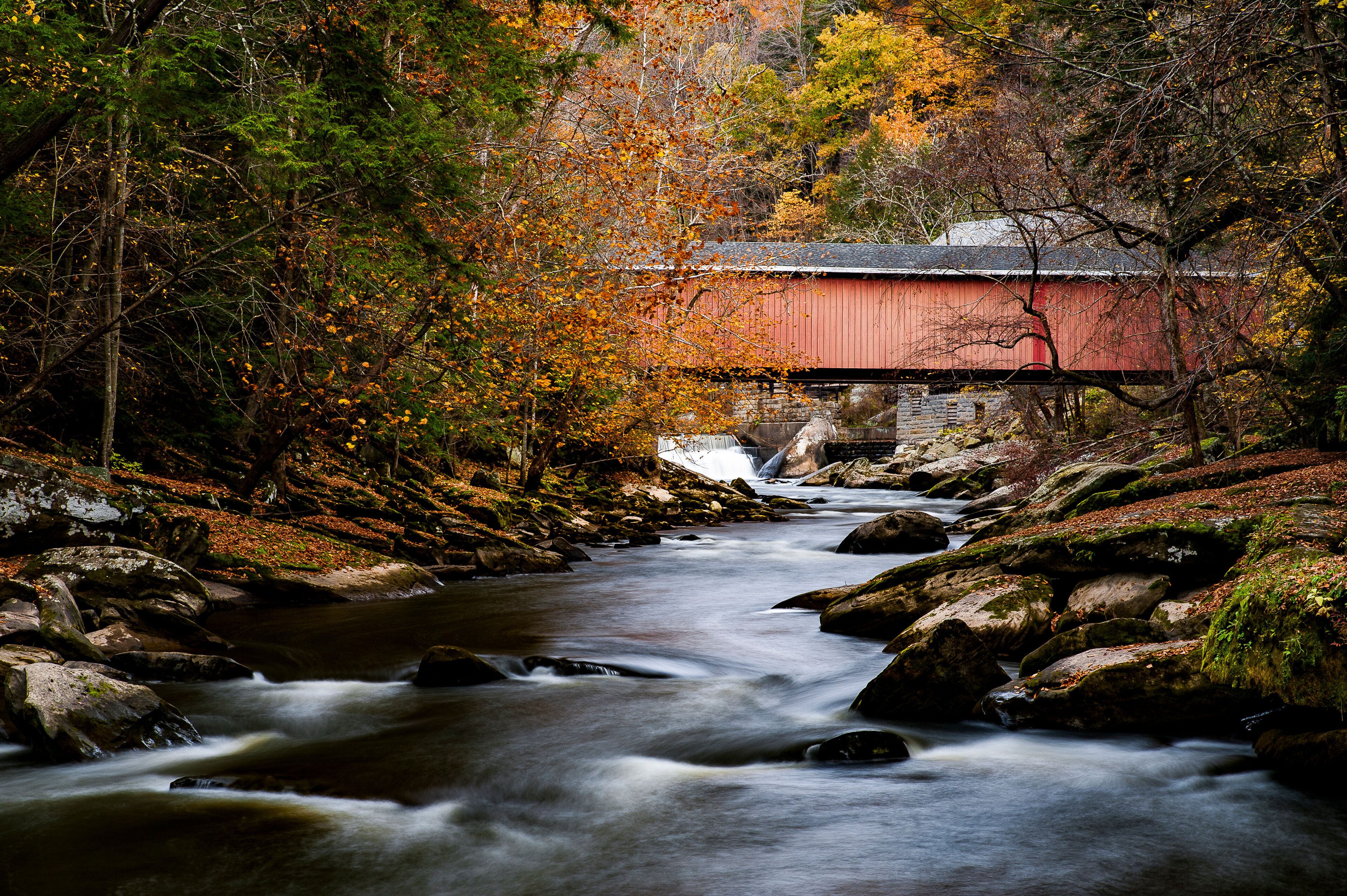 A red covered bridge over a flowing river, surrounded by autumn foliage and rocky terrain.