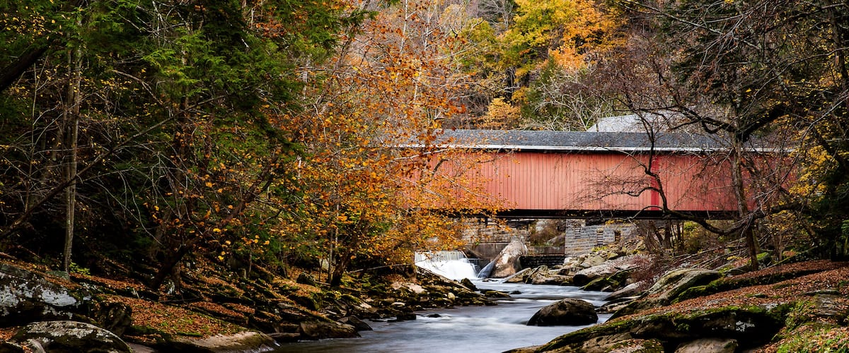 A red covered bridge over a flowing river, surrounded by autumn foliage and rocky terrain.