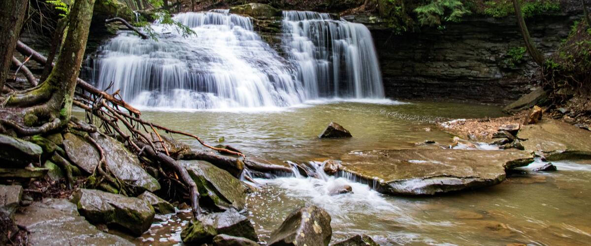 Ein Wasserfall in bewaldeter Landschaft mit felsigem Gelände und üppigem Grün.