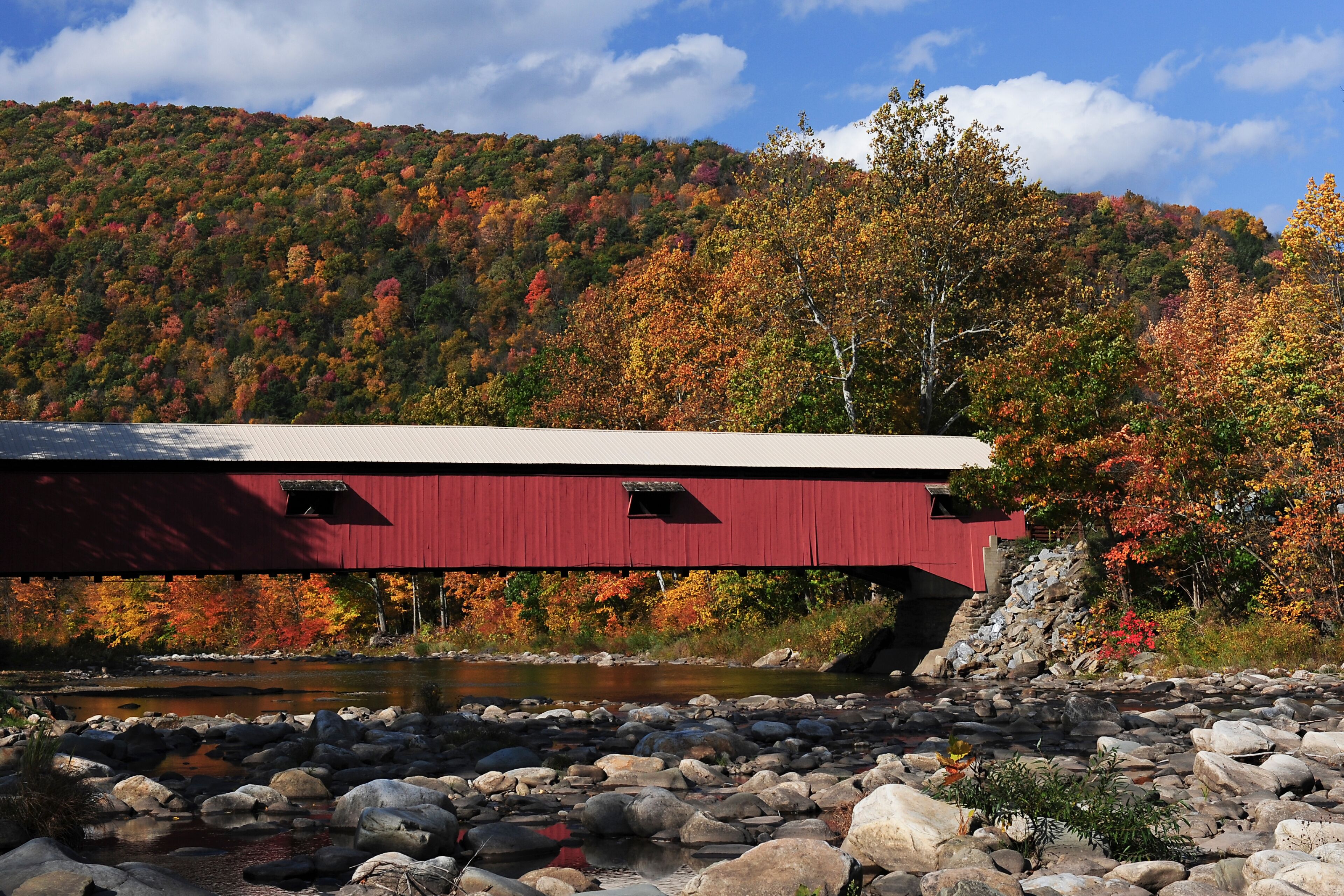 A red covered bridge over a rocky riverbed with autumn foliage in the background.