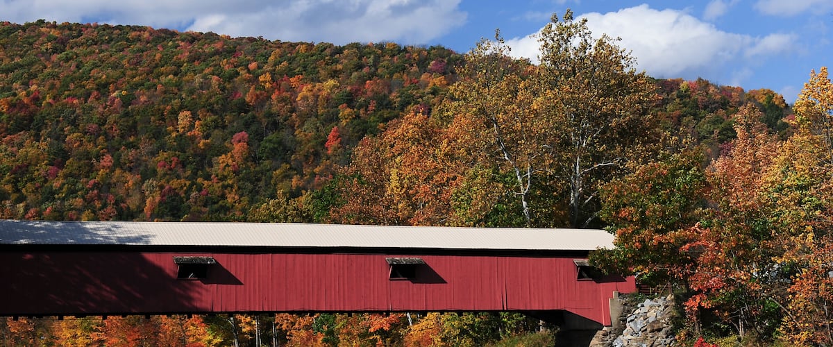 A red covered bridge over a rocky riverbed with autumn foliage in the background.