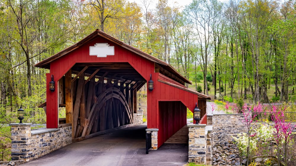 A red covered bridge with stone walls, surrounded by trees and blooming flowers.