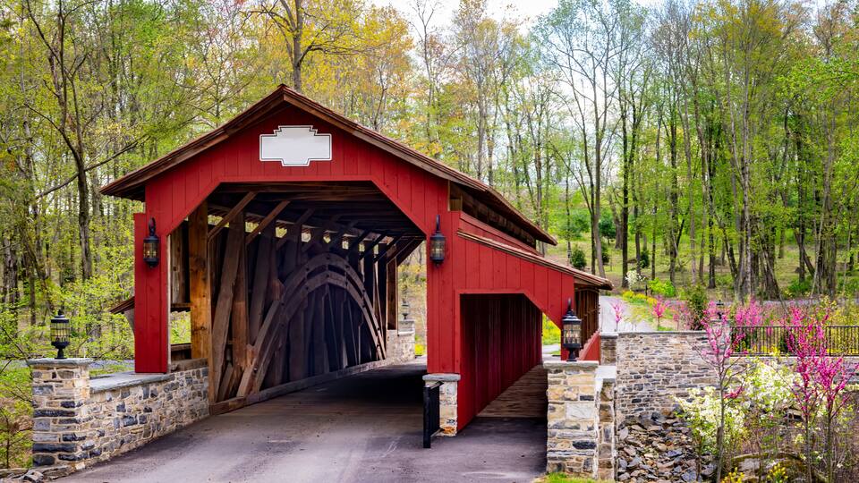 A red covered bridge with stone walls, surrounded by trees and blooming flowers.