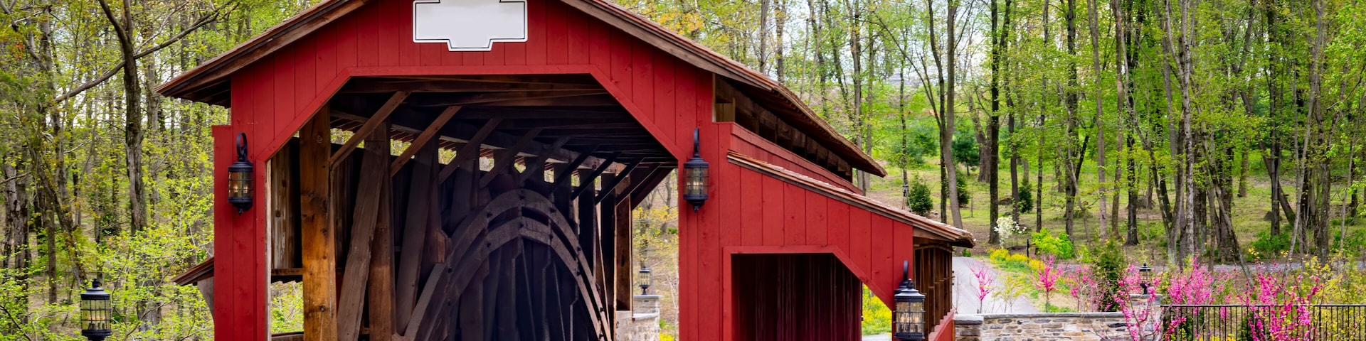 A red covered bridge with stone walls, surrounded by trees and blooming flowers.