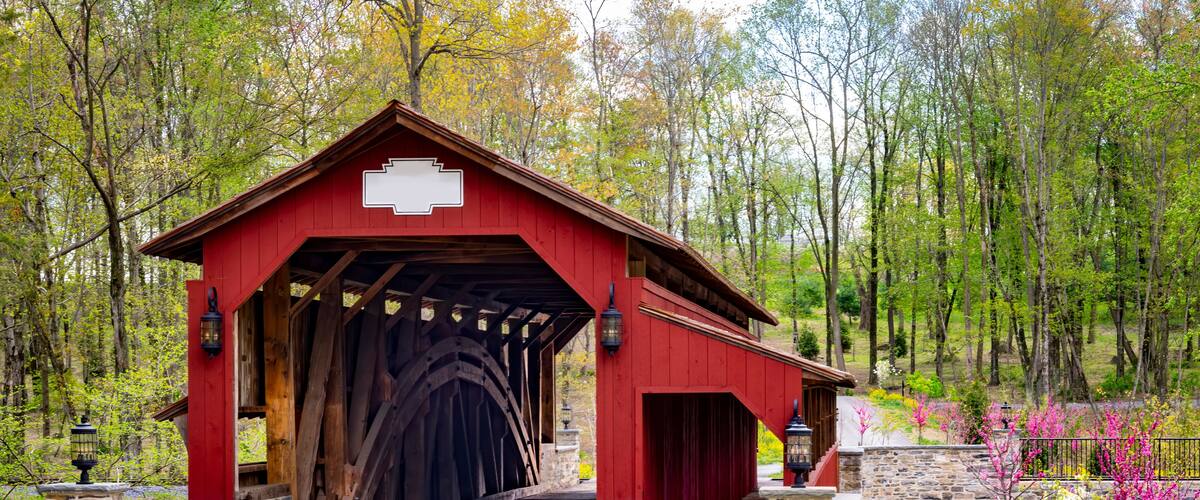 A red covered bridge with stone walls, surrounded by trees and blooming flowers.