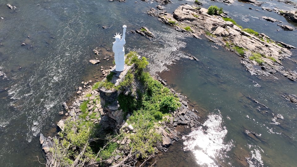 A rocky outcrop with a statue of a figure, surrounded by clear water and greenery.