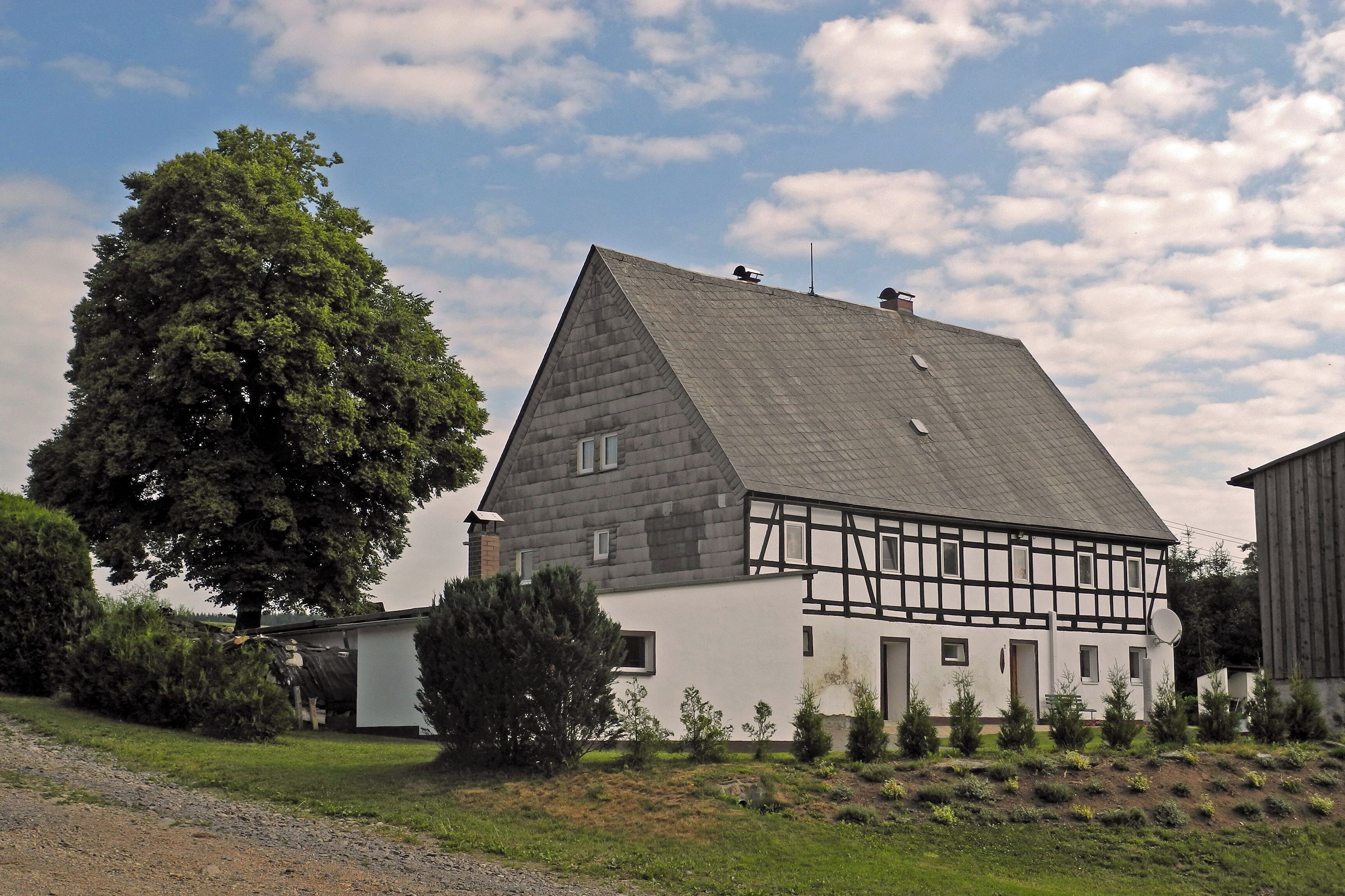 A traditional half-timbered house with a sloped roof, surrounded by a well-maintained garden and a large tree.