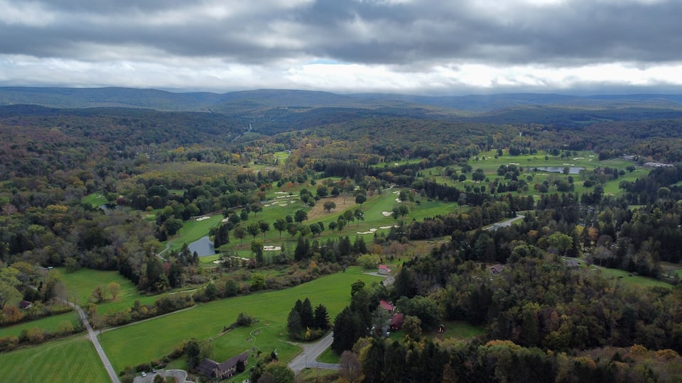 A golf course surrounded by forested hills and a cloudy sky.