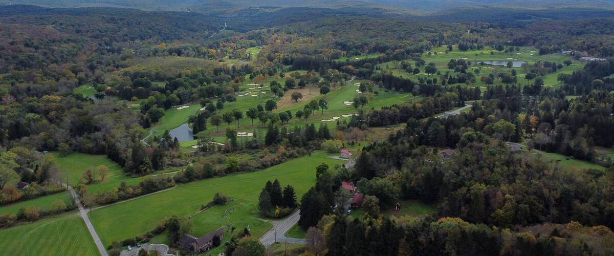 A golf course surrounded by forested hills and a cloudy sky.