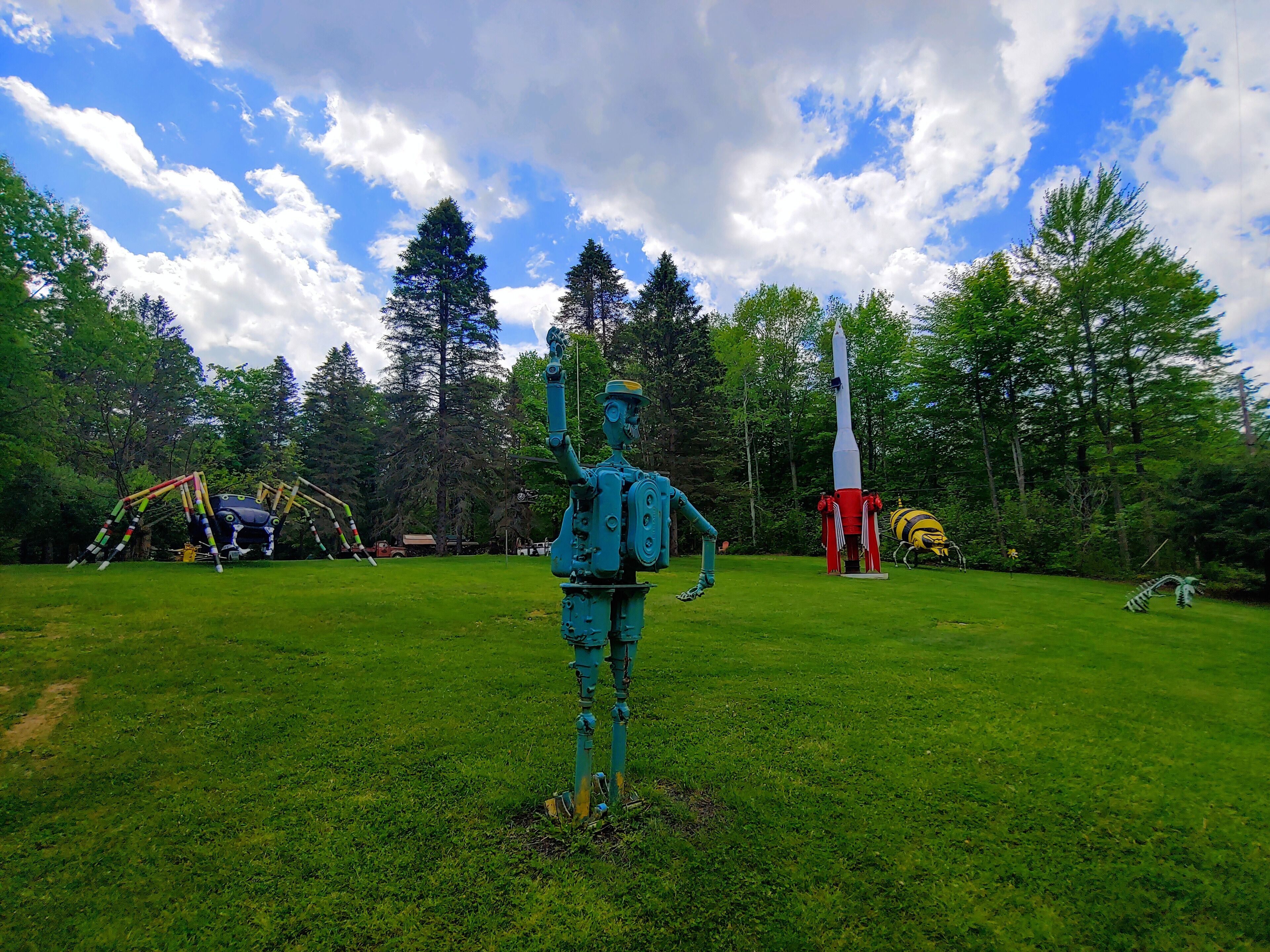 A sculpture of a figure in a green suit with a rocket on a grassy field, surrounded by trees and a rocket launch pad.