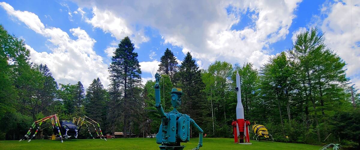 A sculpture of a figure in a green suit with a rocket on a grassy field, surrounded by trees and a rocket launch pad.