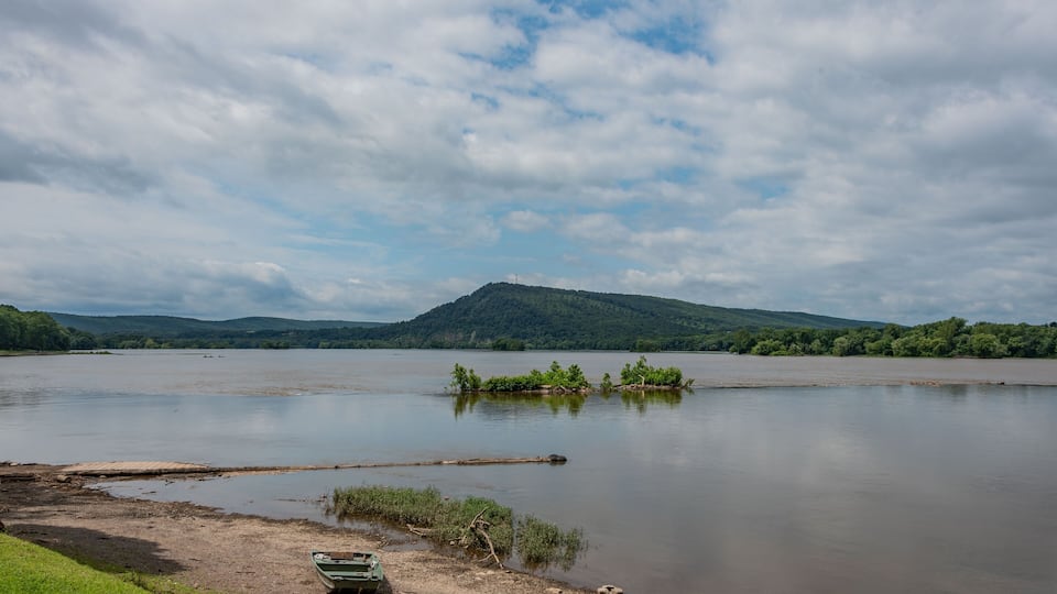 A calm river with a small boat on the shore, surrounded by greenery and hills in the background.