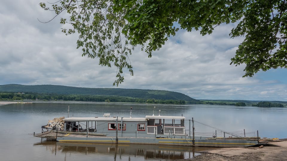 A riverboat docked at a calm river with a forested hillside in the background.