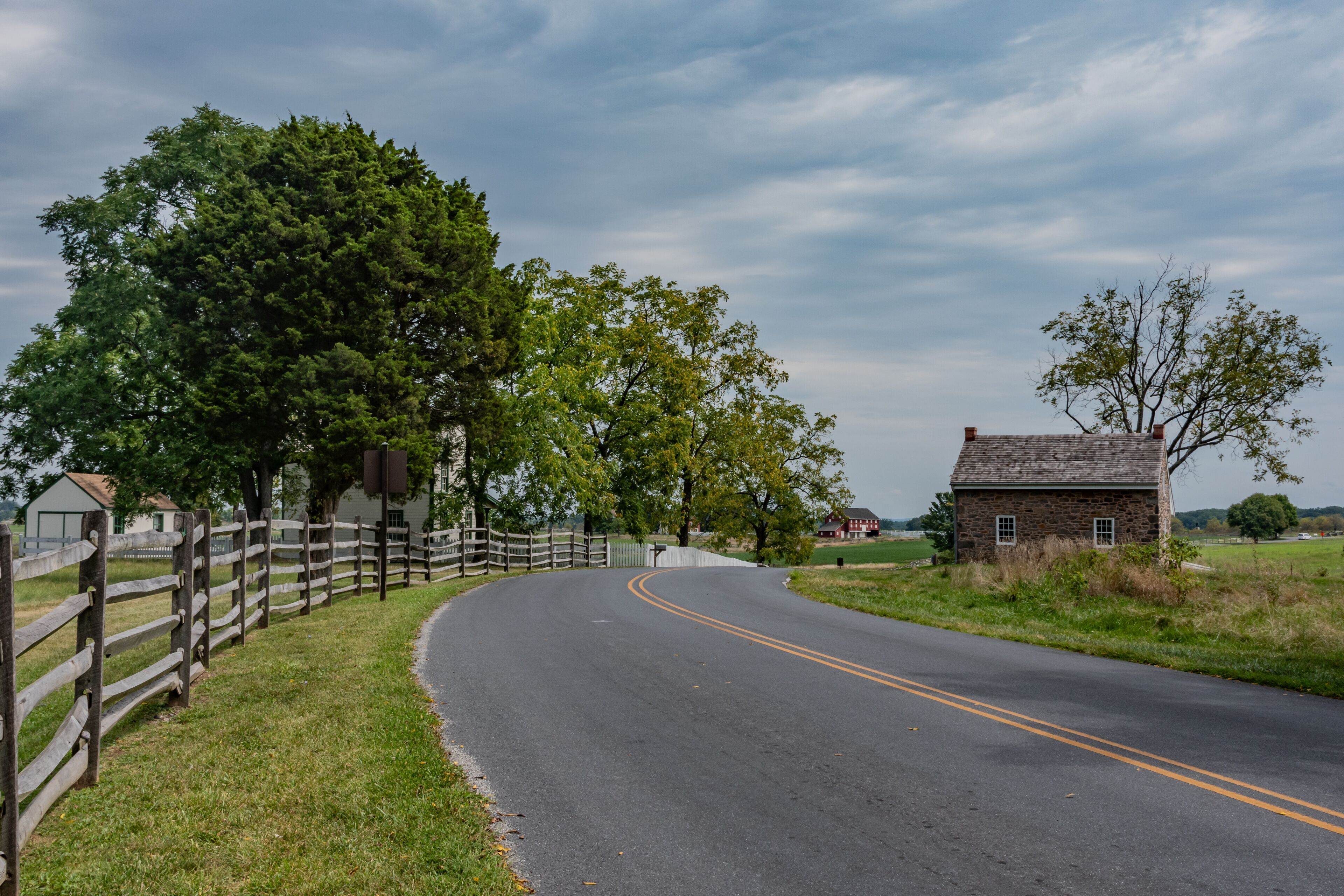 A rural road curving through a landscape with a wooden fence, a stone house, and lush greenery.