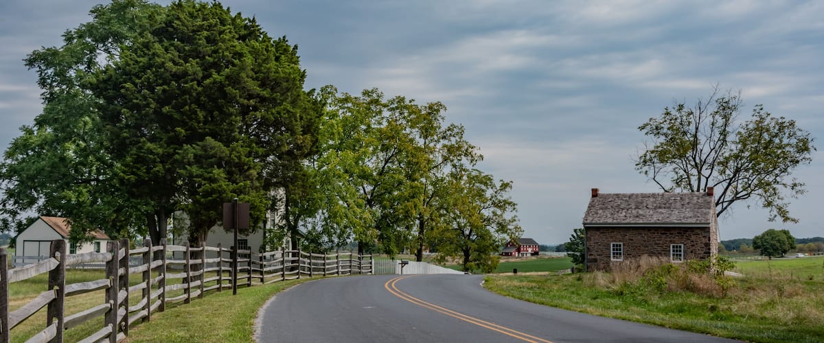 A rural road curving through a landscape with a wooden fence, a stone house, and lush greenery.