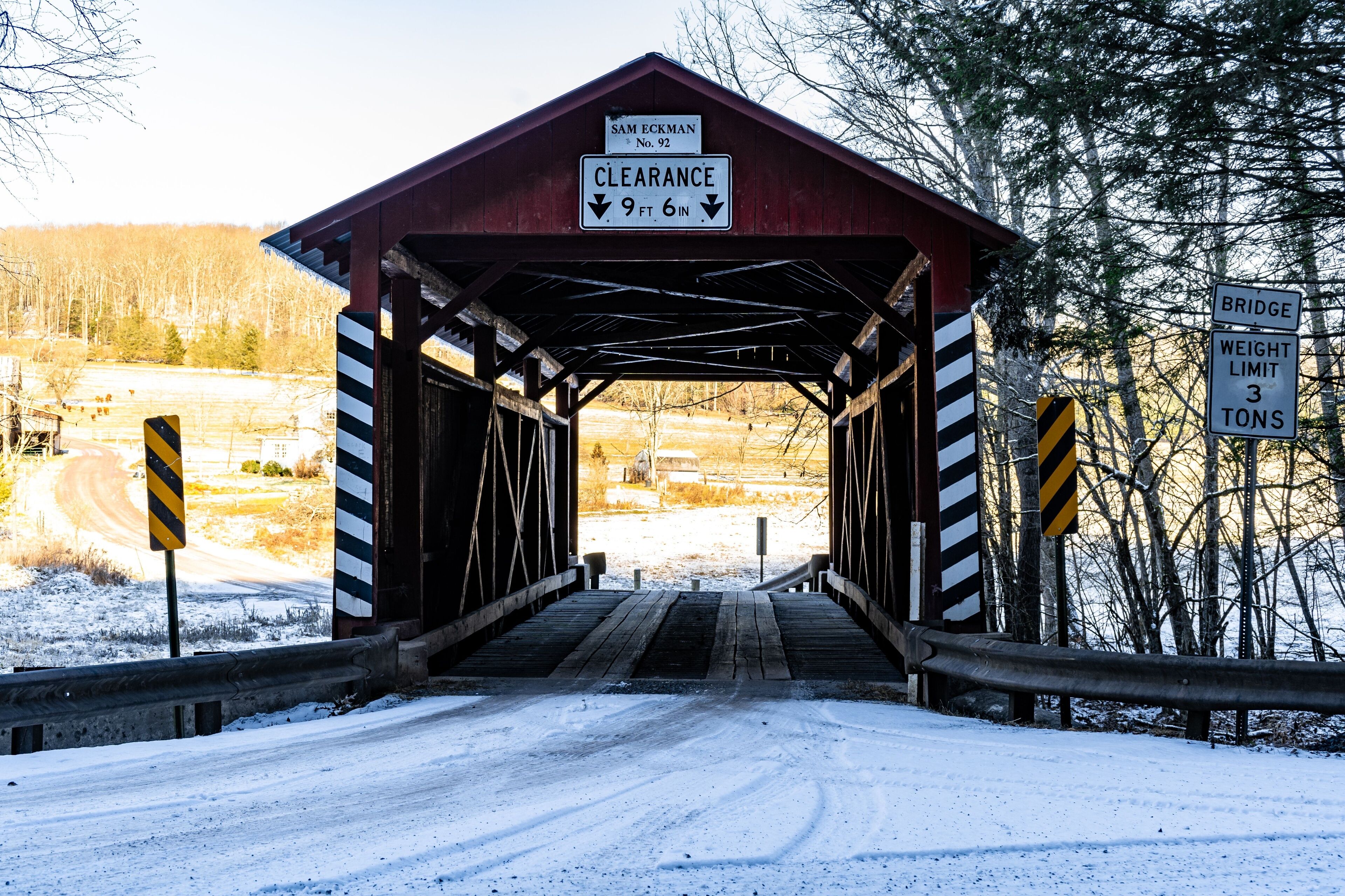 A covered bridge with a clearance sign of 9 ft 6 in, a bridge weight limit sign of 3 tons, and a road leading through a snowy landscape.