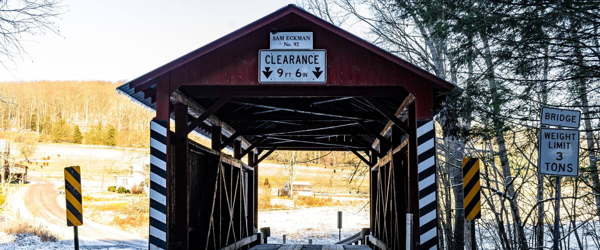 A covered bridge with a clearance sign of 9 ft 6 in, a bridge weight limit sign of 3 tons, and a road leading through a snowy landscape.