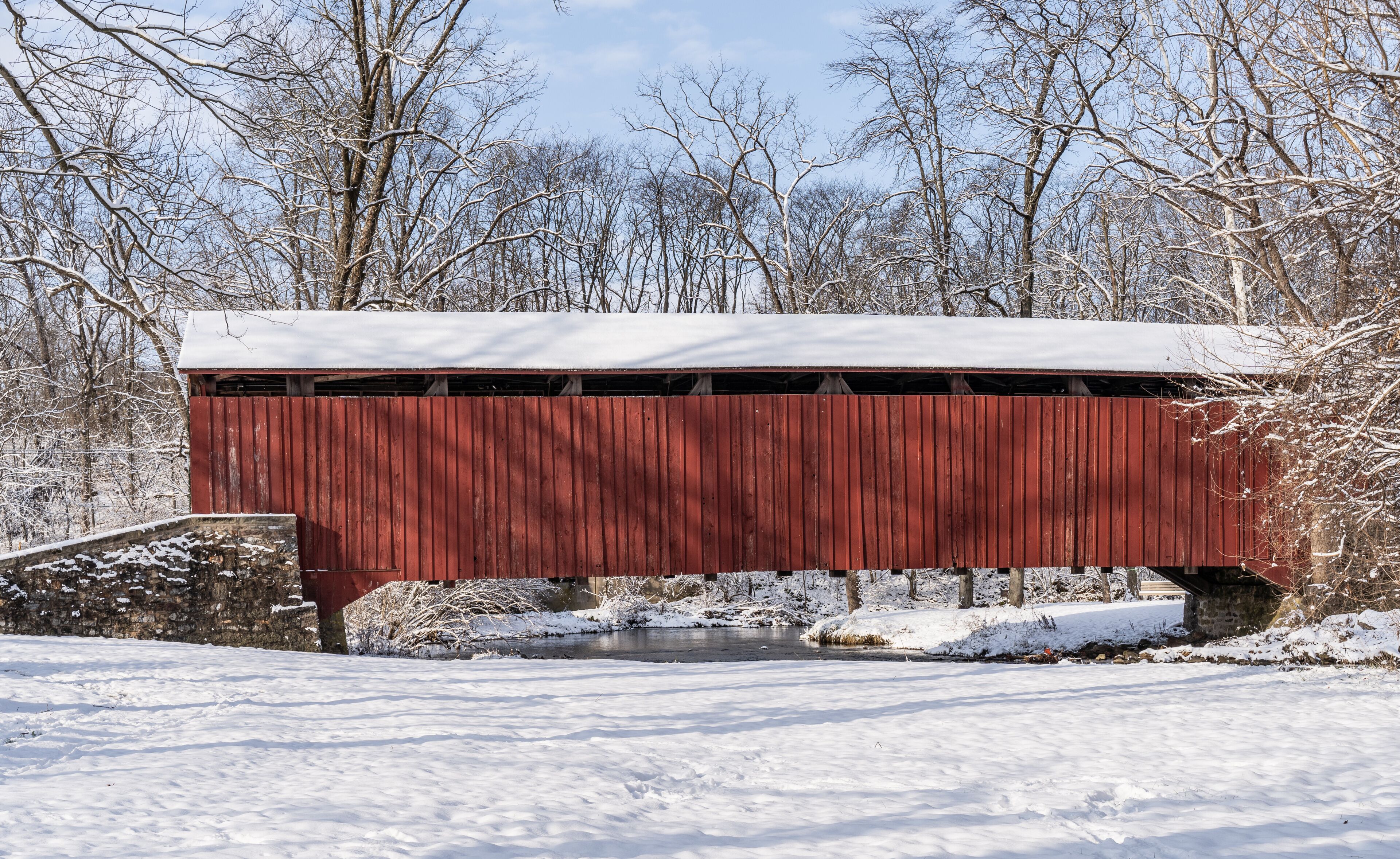 A red covered bridge over a small stream with snow-covered ground and trees in the background.