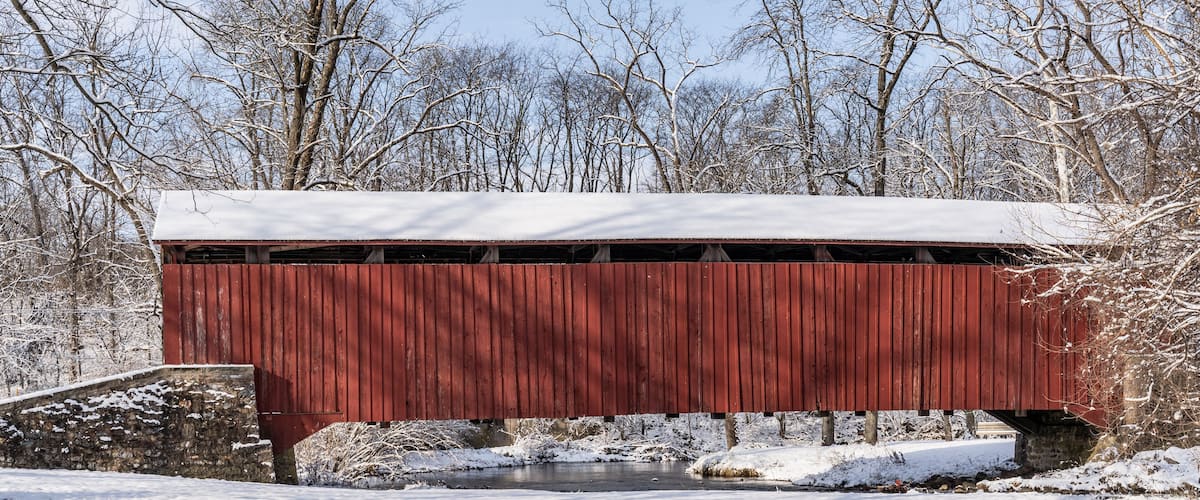 A red covered bridge over a small stream with snow-covered ground and trees in the background.