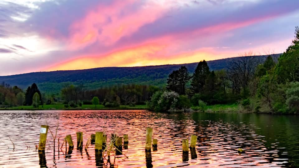 A sunset over a lake with wooden posts in the water, surrounded by trees and hills.