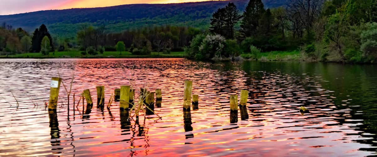 A sunset over a lake with wooden posts in the water, surrounded by trees and hills.