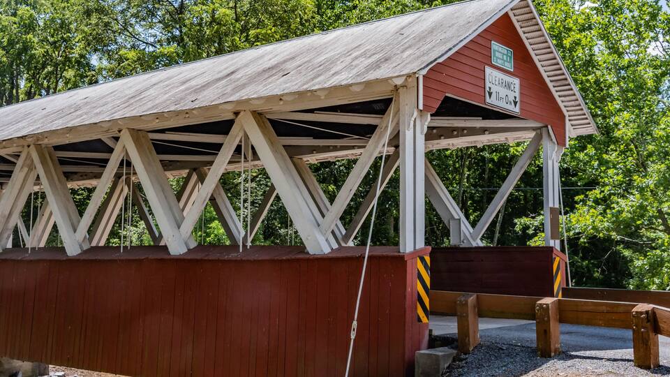 A wooden covered bridge with a red roof and white beams, surrounded by trees.
