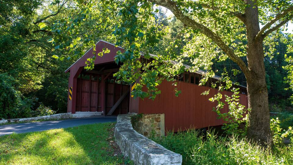 A red covered bridge with stone walls, surrounded by lush green trees and grass.