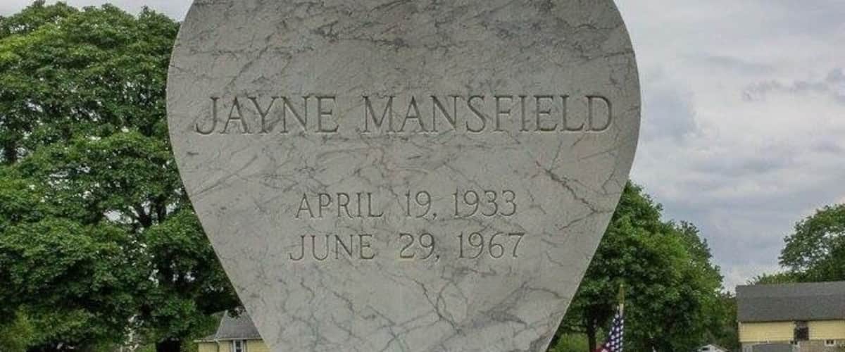 A heart-shaped gravestone with Jayne Mansfield's name and dates, surrounded by American flags.